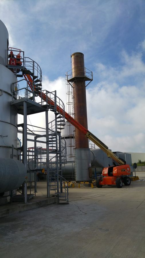 A boom lift working alongside tanks and a chimney stack at an industrial plant