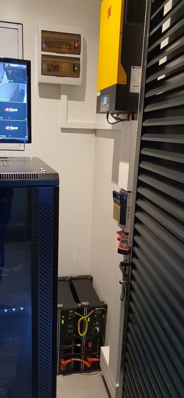 A wall-mounted inverter and distribution boards above a battery bank beside a server cabinet